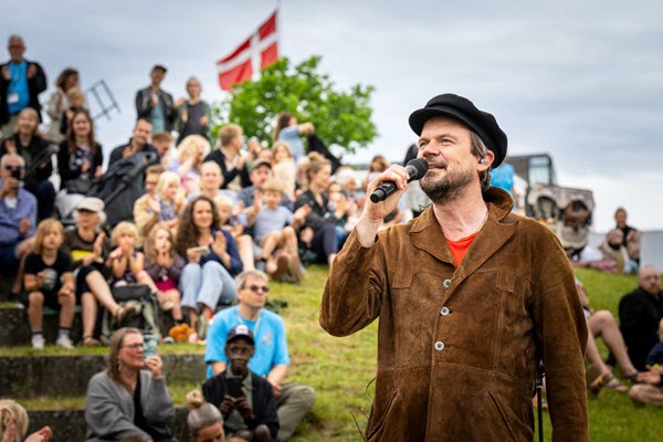 Peter Sommer optræder foran et engageret udendørs publikum ved Sløngeldagene, med Dannebrog vajende i baggrunden.